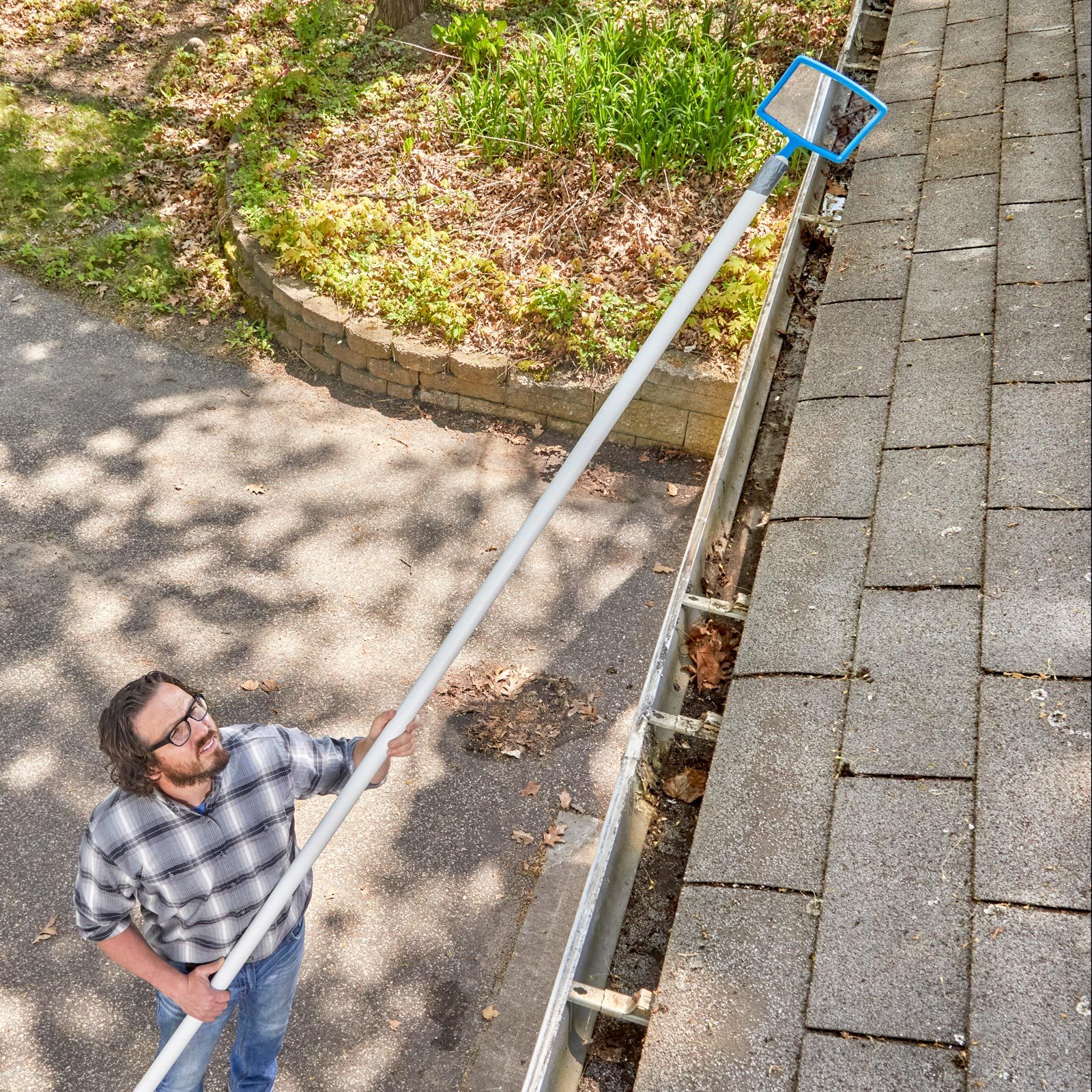 A man uses a long pole with a scoop to clean leaves from a gutter while standing on a paved surface near greenery.