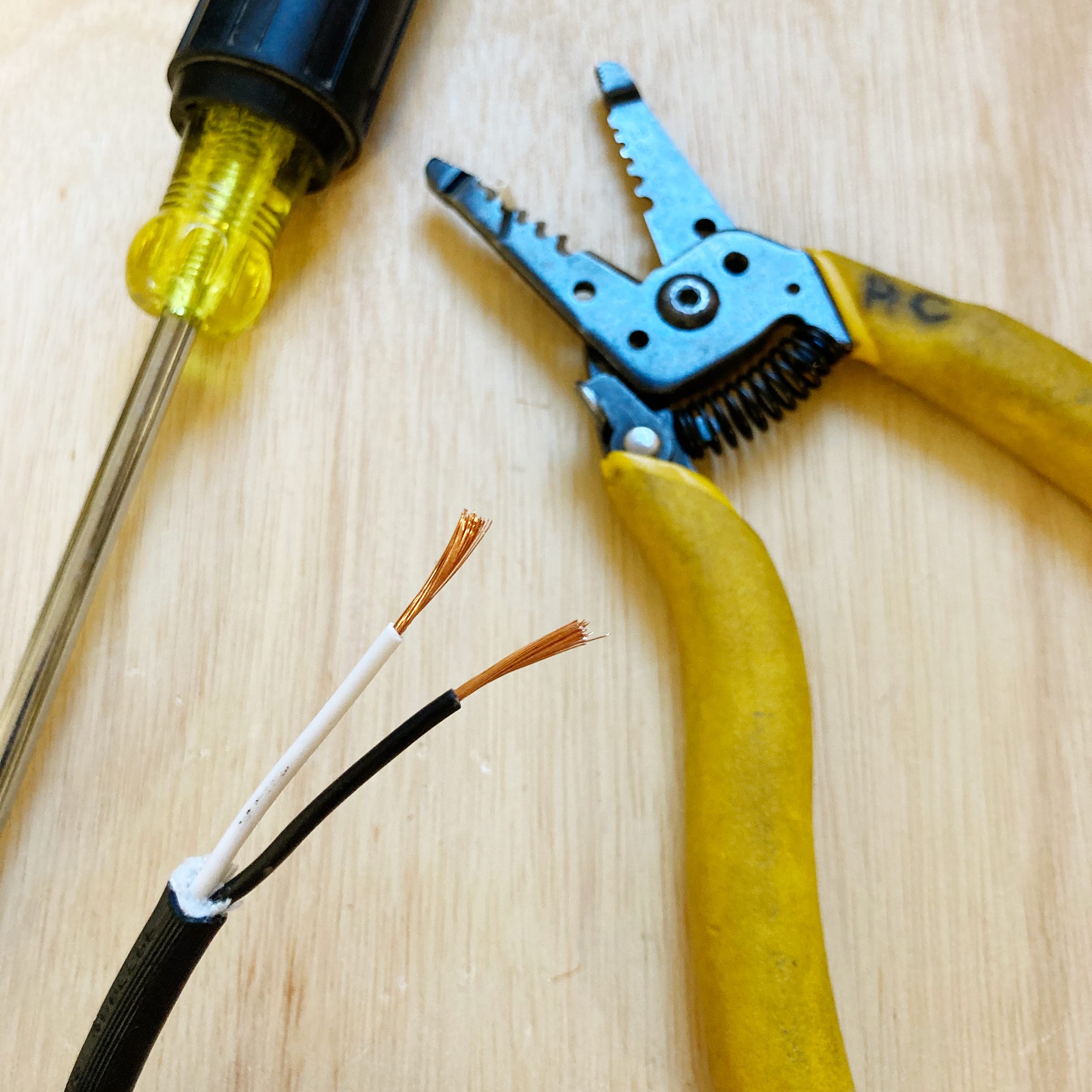 Electrical wires with exposed copper ends sit on a wooden surface, while a pair of wire strippers and a yellow screwdriver are nearby, suggesting preparation for electrical work.