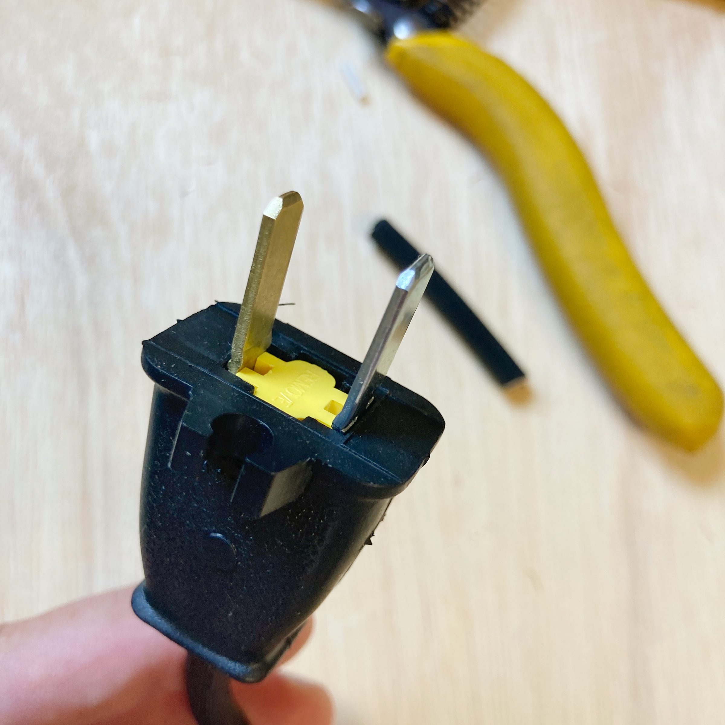 A person holds a black electrical plug with two metal prongs, surrounded by tools and a banana on a wooden surface.