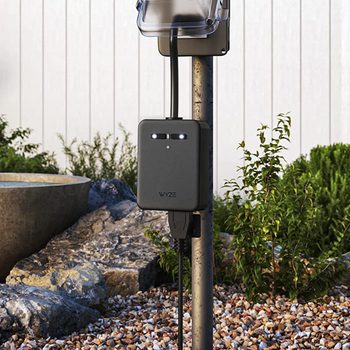 A black electrical box with lights is connected to a pole and outlet, situated among stones and plants near a water feature in a garden setting.