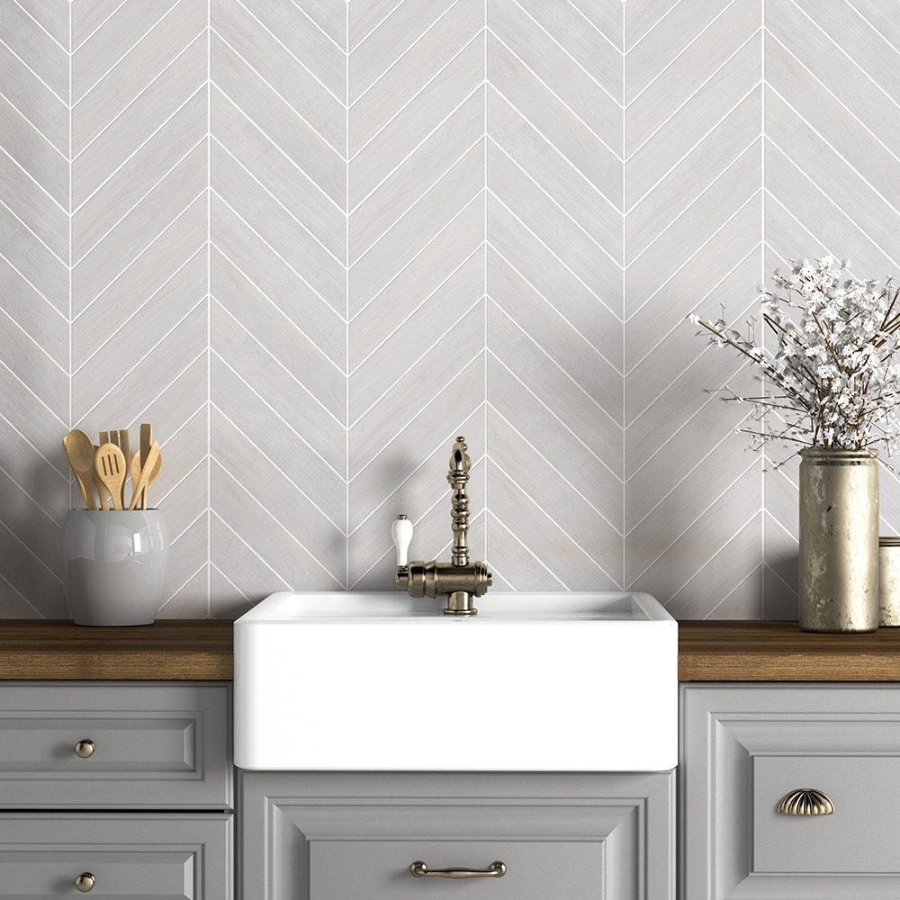 A white sink sits on a wooden countertop, with a vintage faucet. Gray chevron-patterned walls surround kitchen utensils and a decorative vase with branches.