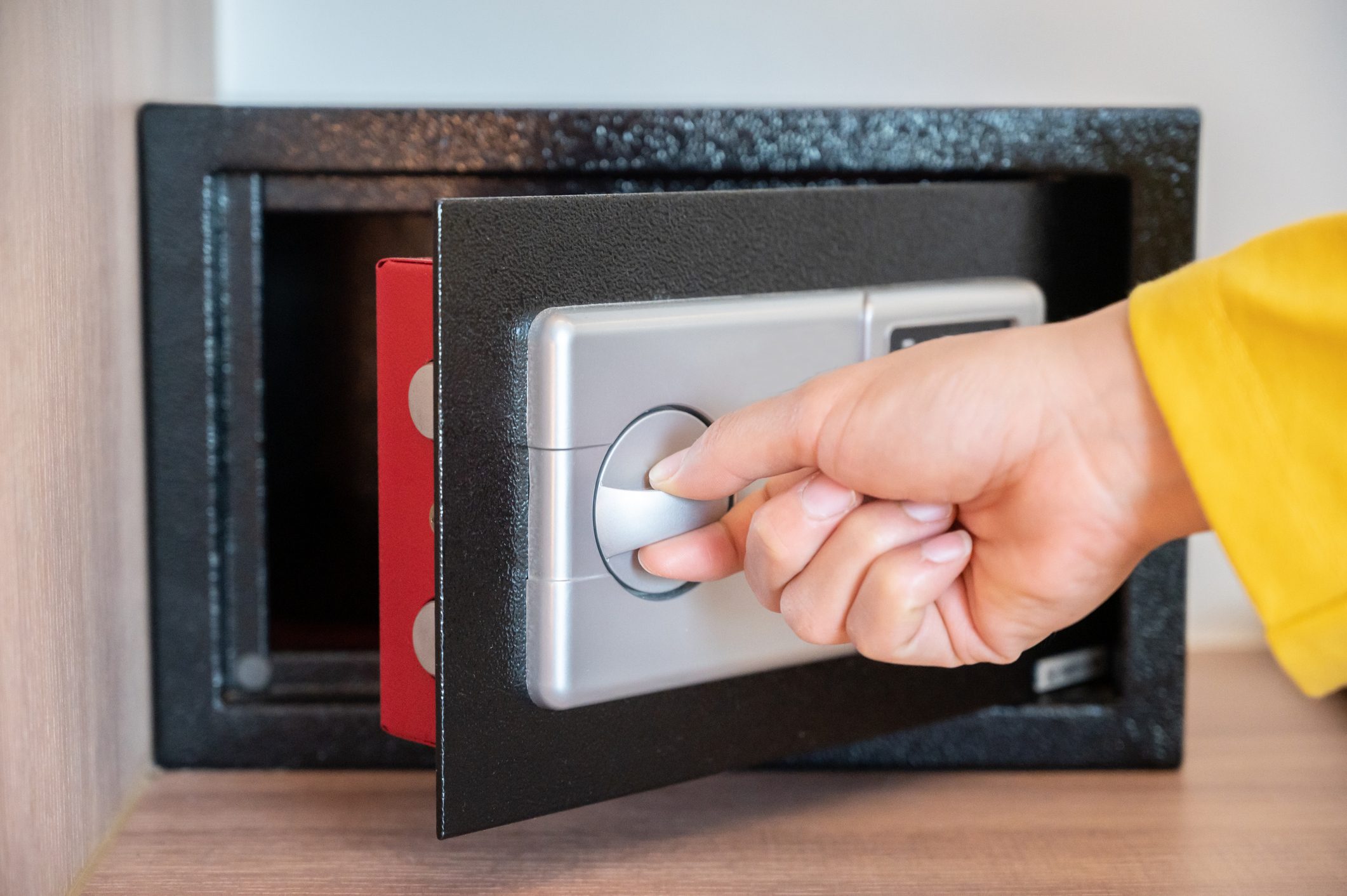 A hand turns a lock on a black safe, which is partially open, revealing a red item inside on a wooden surface.