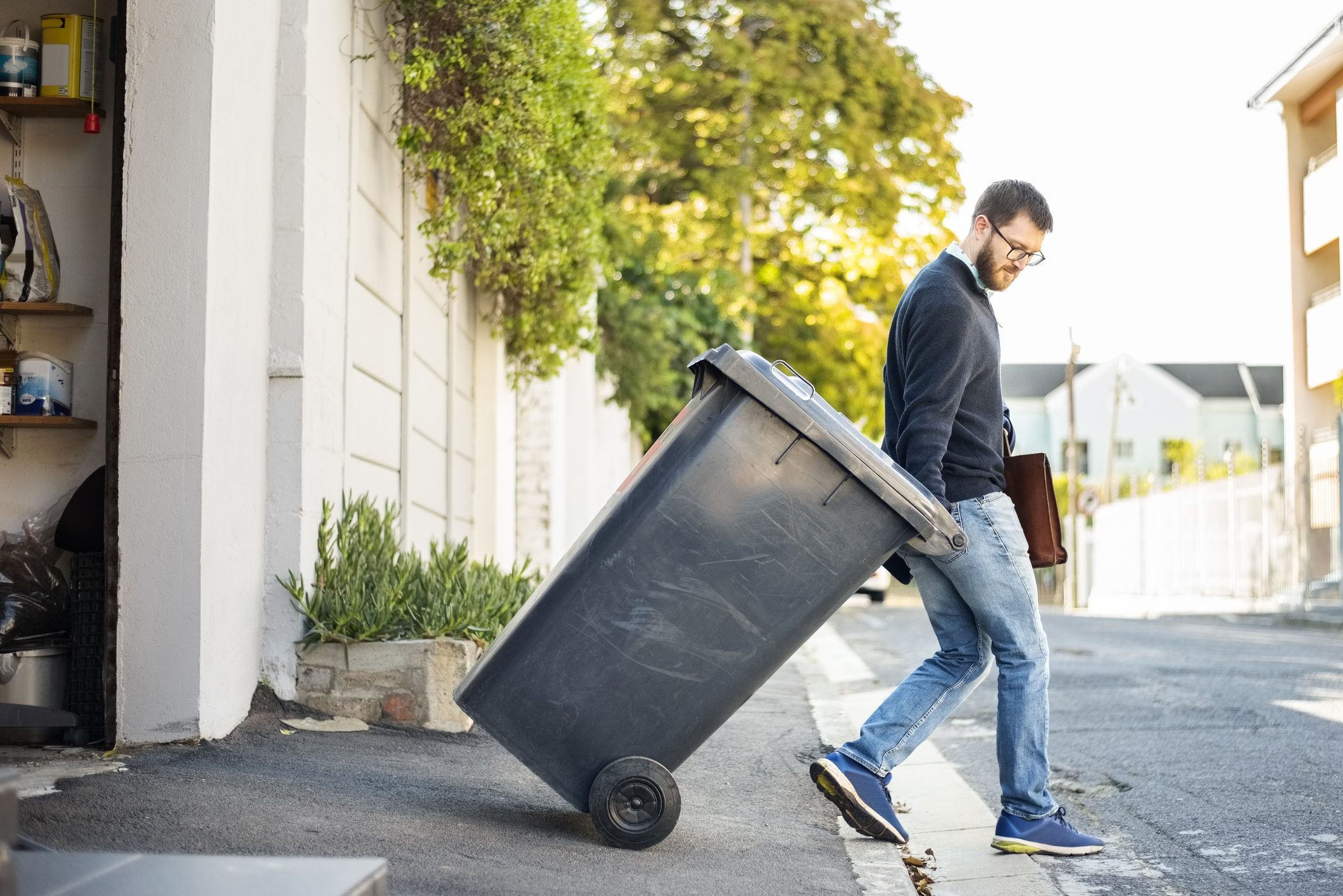 A man pushes a large trash bin along a sidewalk, surrounded by greenery and residential buildings under clear skies.