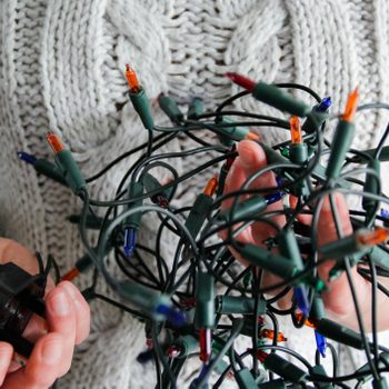 A person holds a tangled mass of colorful Christmas lights, with a plug in one hand, wearing a cozy, textured sweater.