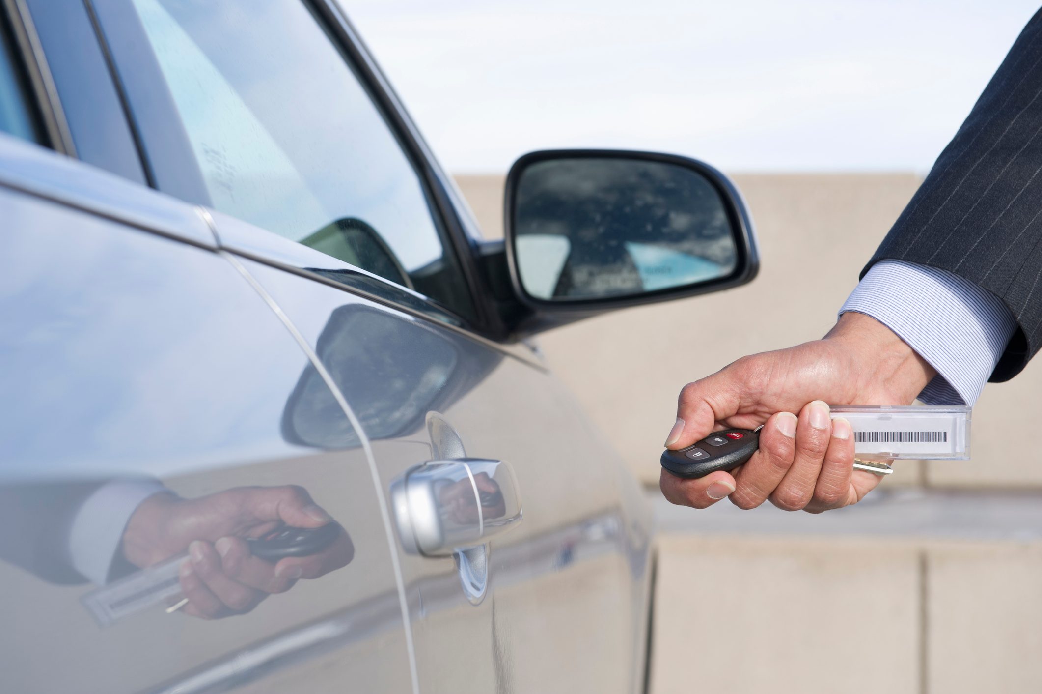 A hand holds a car key fob near a silver car door, preparing to unlock it against a background of a clear sky and concrete.