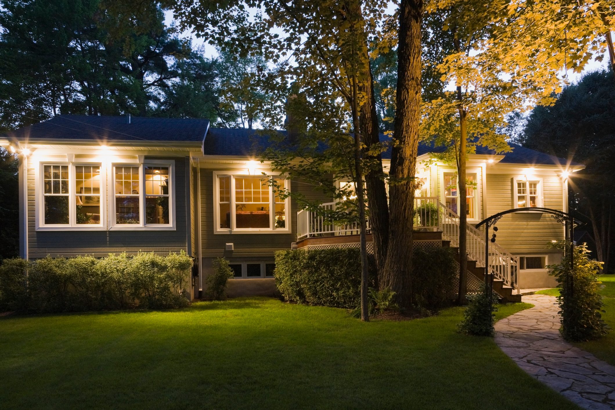 A well-lit home with large windows stands in a lush lawn, surrounded by trees, evoking a cozy atmosphere during twilight.