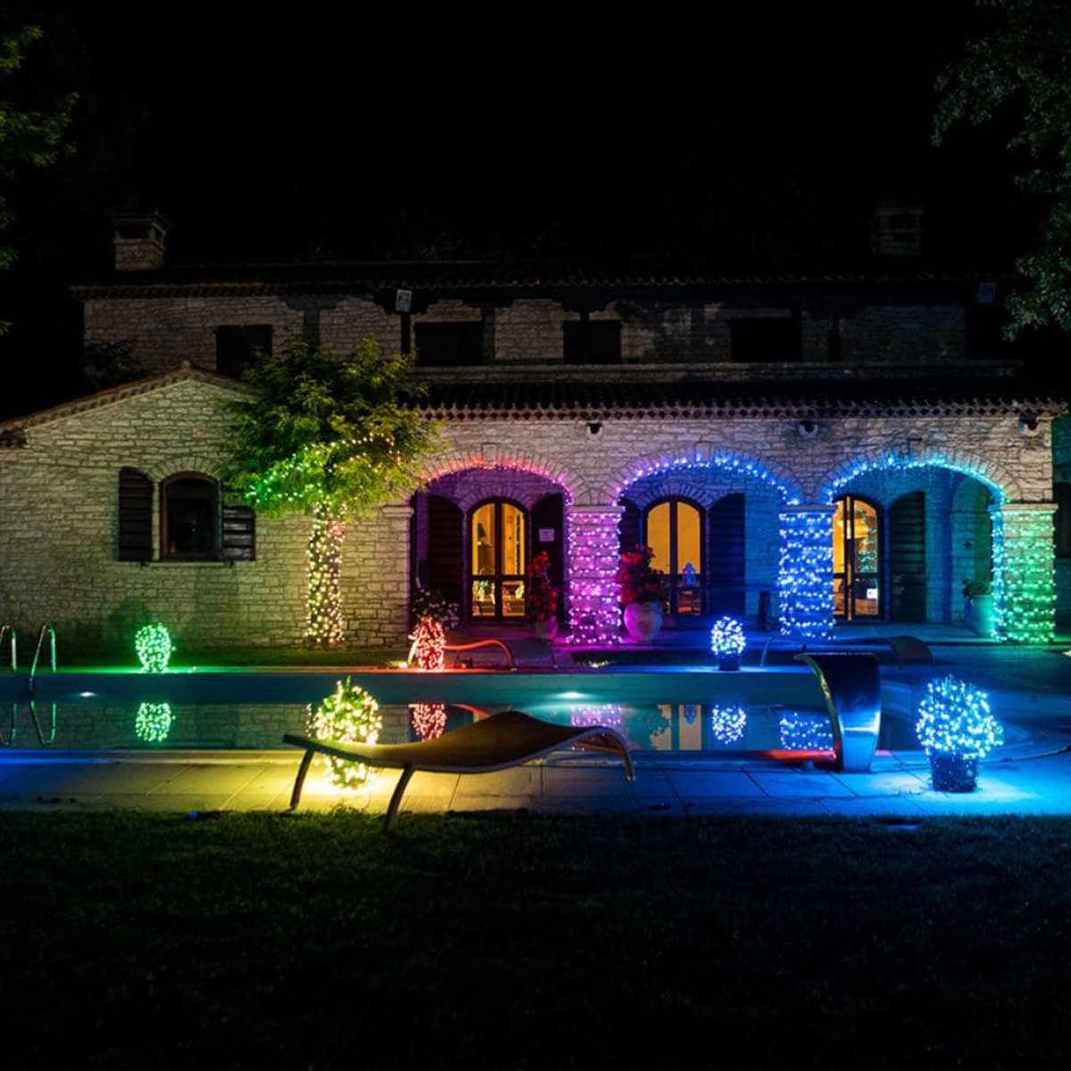 A stone house illuminated with colorful lights surrounds a pool, reflecting vibrant hues. Lounge chairs sit beside the water in a serene nighttime setting.