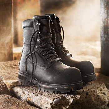 Black work boots rest on a textured, rocky surface, surrounded by a misty, industrial environment, suggesting rugged use and durability.