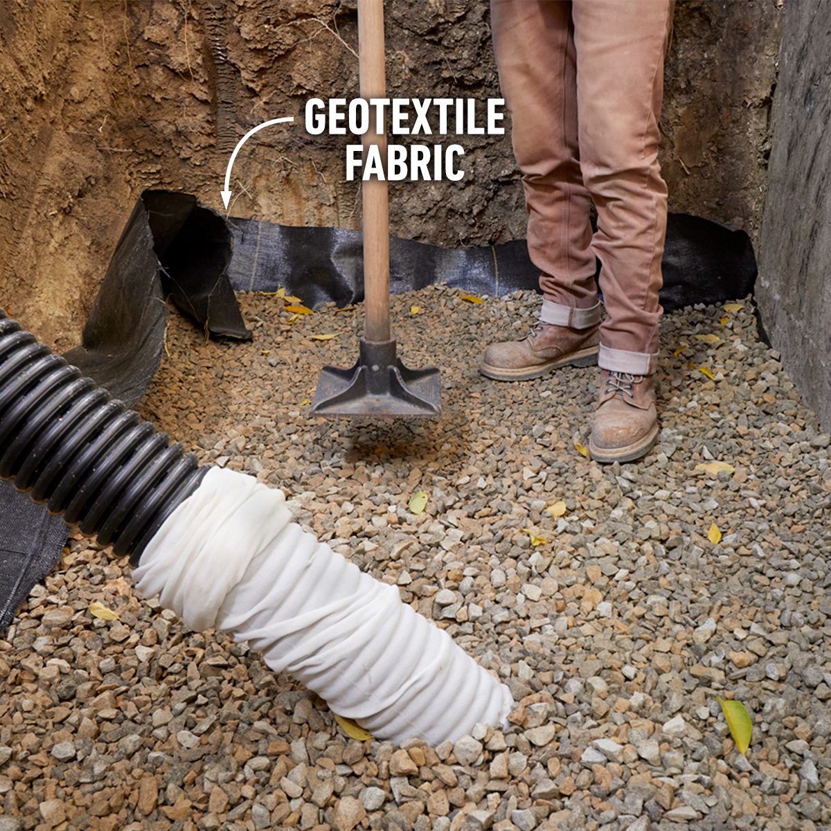 A person stands on gravel, holding a shovel next to a black drainage pipe covered with white fabric, beside visible geotextile fabric.