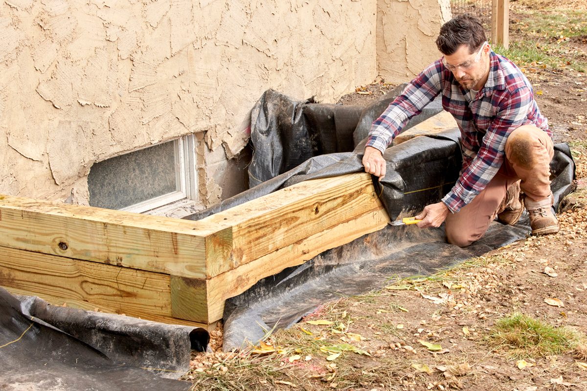 A man is measuring and positioning wooden planks against a house's foundation, working in a garden area with some soil and leaves around.