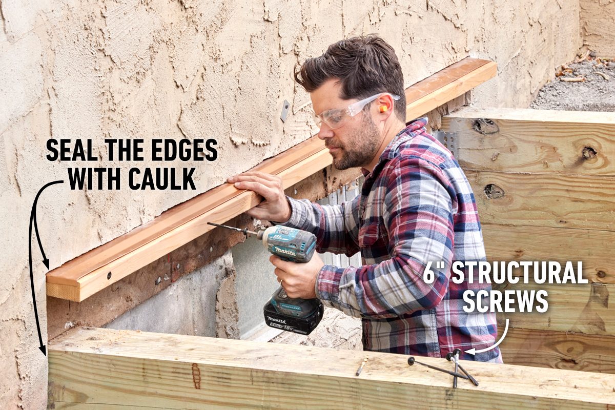 A man drills screws into a wooden beam, securing it against a textured wall while preparing to seal edges with caulk in a construction setting.