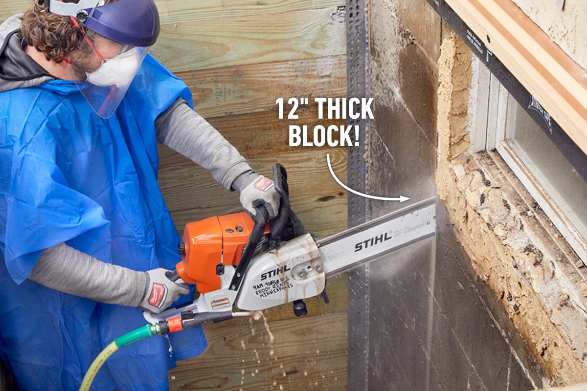 A person in protective gear operates a chainsaw, cutting through a thick block wall, while dust and debris are expelled in a construction environment.
