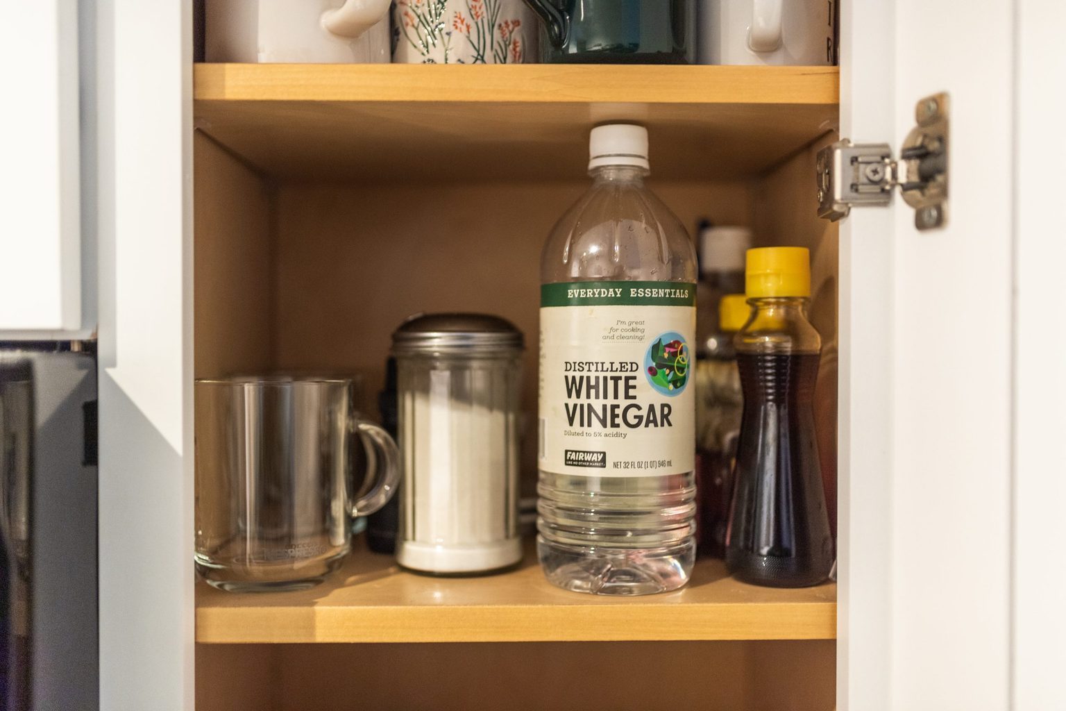 A bottle of distilled white vinegar stands among various cooking ingredients on a wooden shelf, alongside a glass mug and a sugar container.