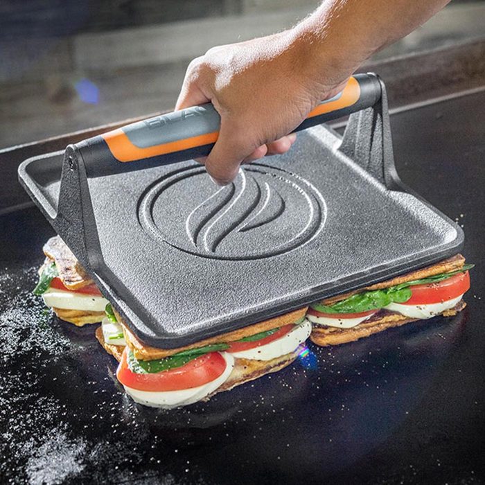 A hand lifts a heavy, ridged press above a sandwich with layers of vegetables and cheese on a griddle, indicating the cooking process.