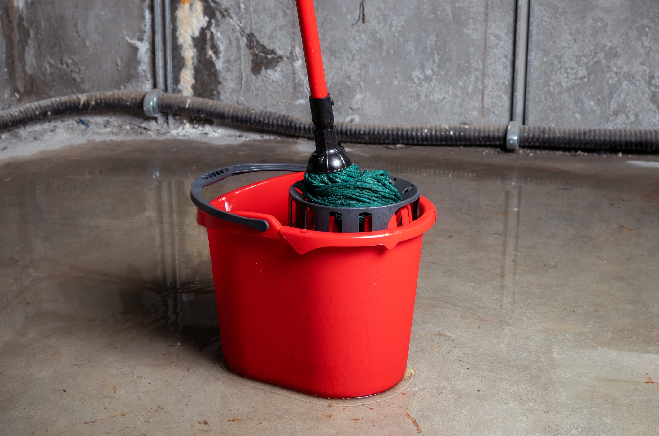 A red bucket with a mop inside stands on wet concrete. The environment appears unfinished, with exposed walls and plumbing in the background.
