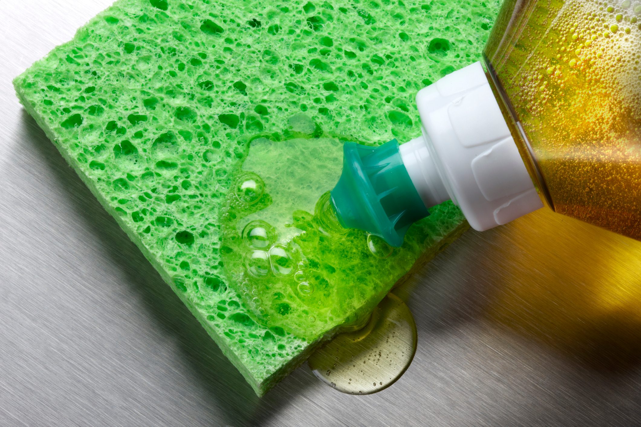 A green sponge absorbs yellow liquid detergent from a tilted bottle, resting on a metallic surface. Bubbles form as the detergent spreads.