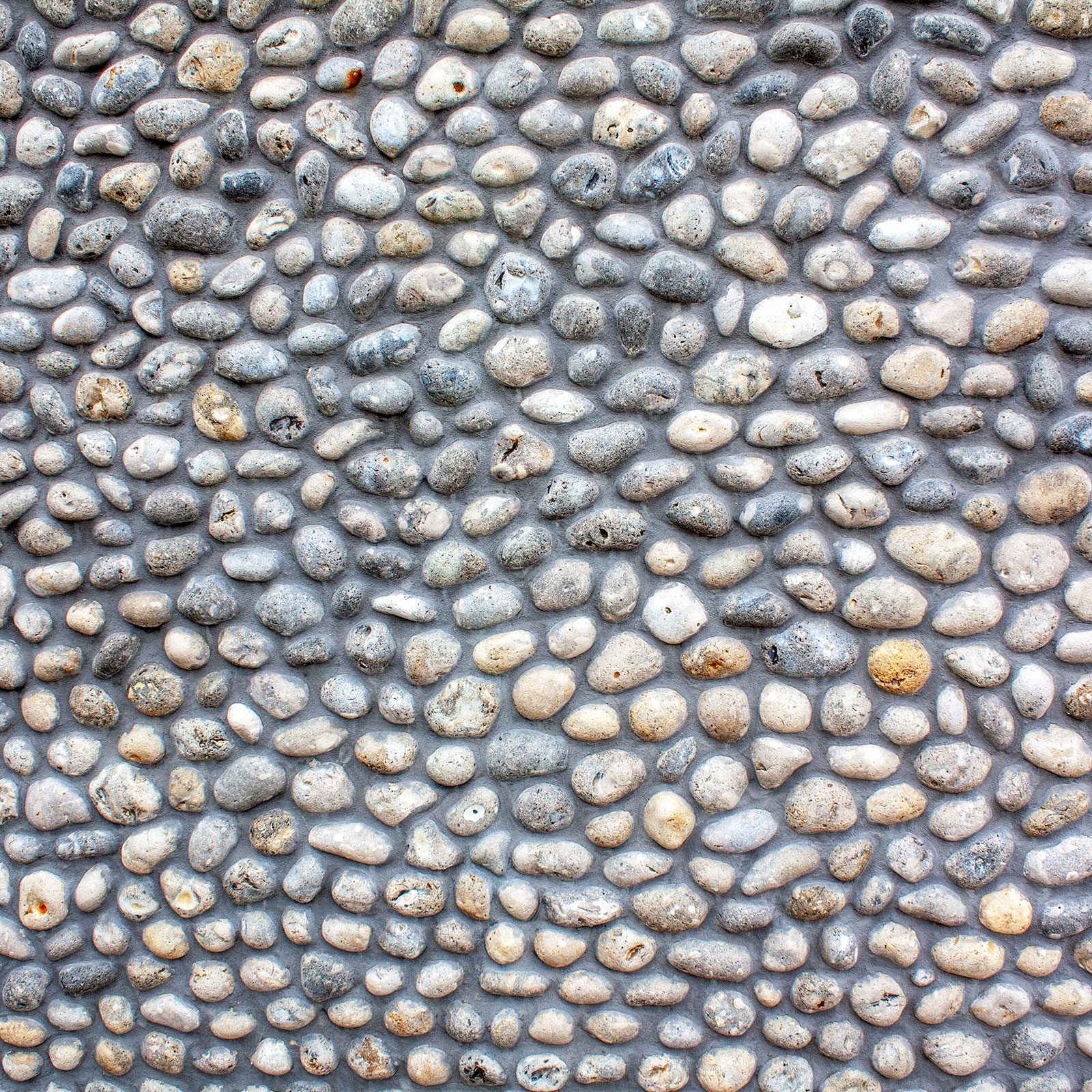 Smooth, rounded pebbles cover a wall, arranged closely together, showcasing various gray and beige tones in a natural, textured pattern.