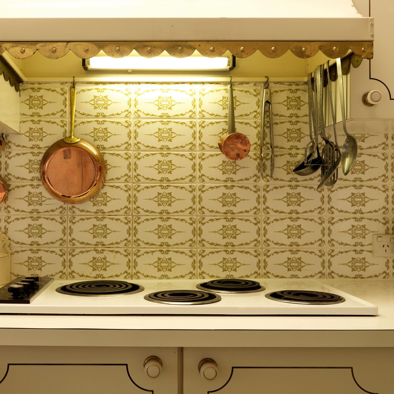 A white stove with circular burners sits below an illuminated hood, surrounded by hanging copper pans and assorted metal utensils against a patterned backsplash.