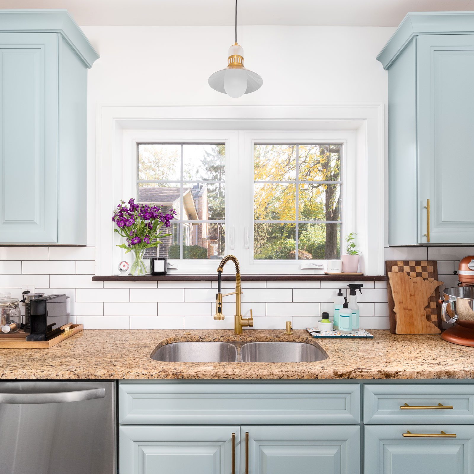 A double sink with a gold faucet sits on a granite countertop, under a window framed by cabinetry, while a potted plant and flowers add color.