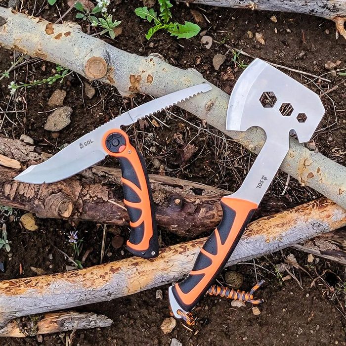Two outdoor tools, a knife and a multi-functional hatchet, rest on a bed of logs and earth, showcasing their metal blades and textured handles.