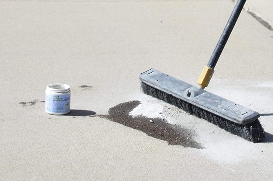 A broom sweeps dirt and debris from a light-colored concrete surface, while a small container sits nearby, possibly containing cleaning material.