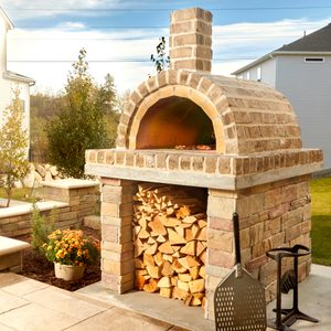 A stone pizza oven sits outdoors, emitting heat while cooking food inside, surrounded by neatly stacked firewood and a landscaped yard.