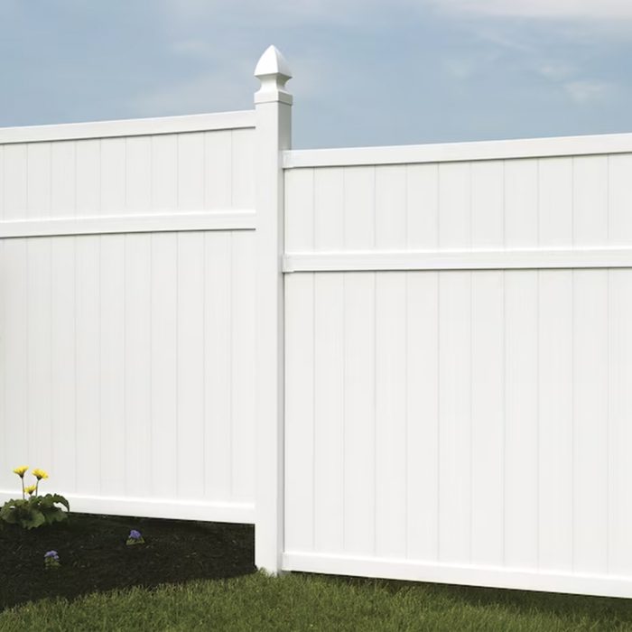 A white vinyl fence segment stands upright, separating areas, with flowers growing at its base and a cloudy sky overhead.