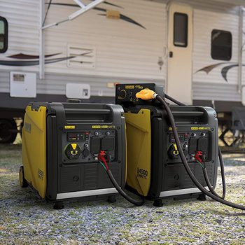Two portable generators are positioned on gravel, connected by a cable, with an RV parked nearby, set in an outdoor setting.