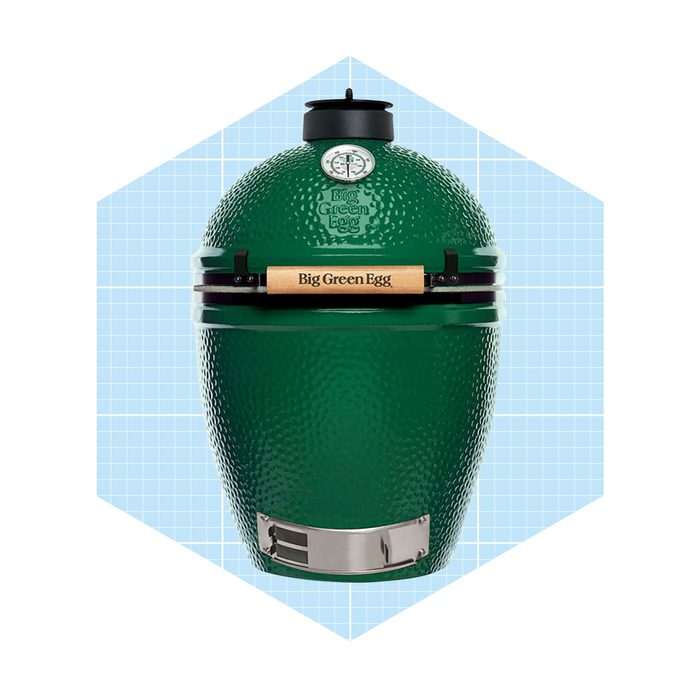 A green, egg-shaped grill sits upright, featuring a wooden handle and metallic bottom. It's set against a light blue, grid-patterned background.