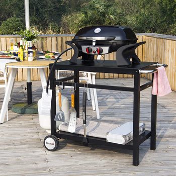 A black gas grill sits on a wheeled cart, with cooking utensils hanging nearby, on a wooden deck surrounded by greenery and a table set for dining.