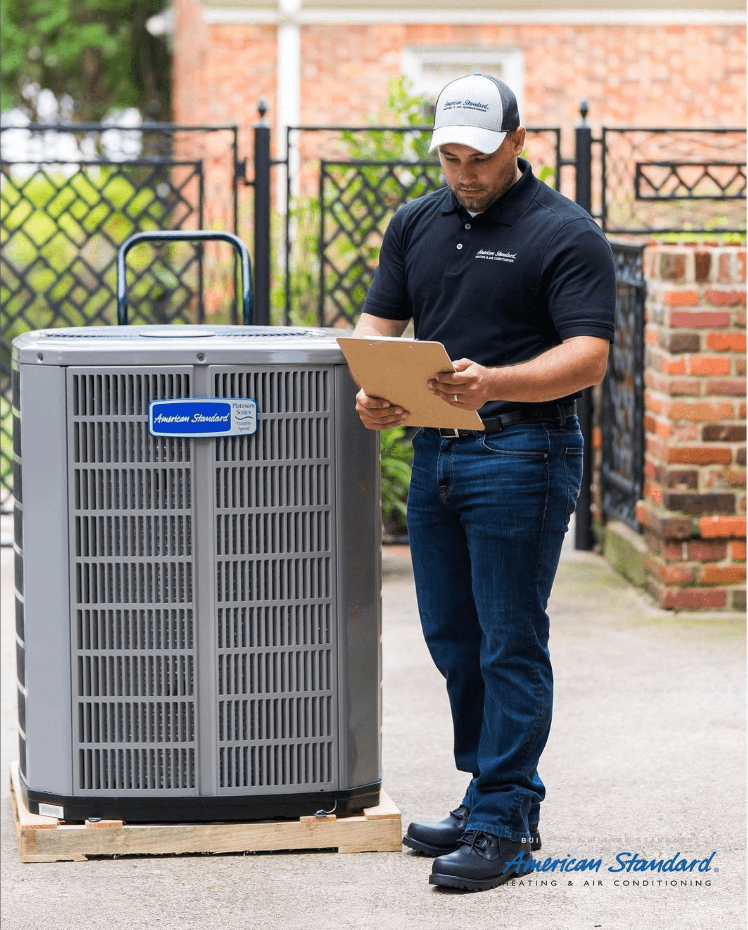 A technician reviews a clipboard while standing next to an air conditioning unit outside, surrounded by a brick wall and green foliage.