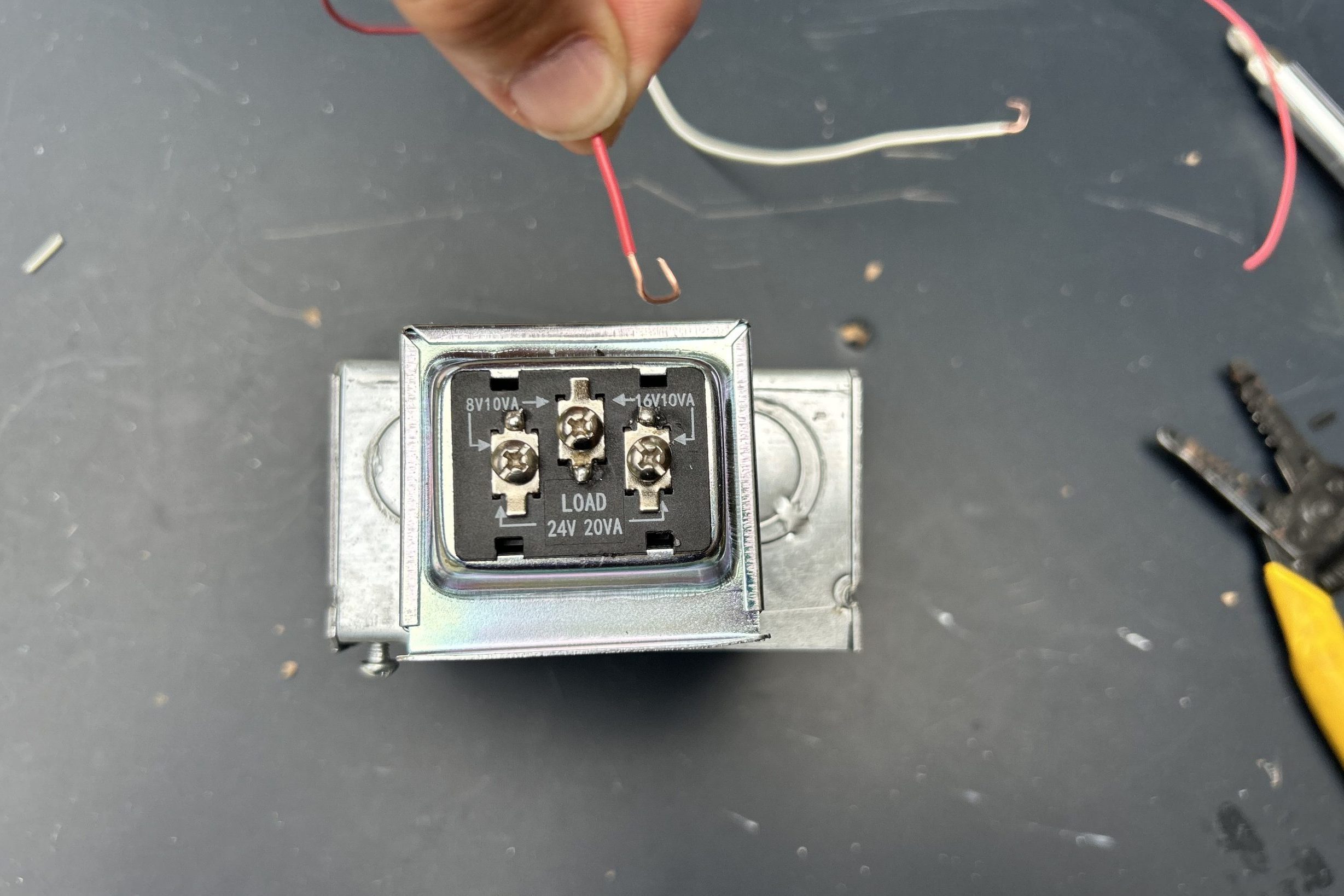 A person holds a red wire near a relay, which has three connection points labeled for voltage. The workspace is cluttered with tools and wires.