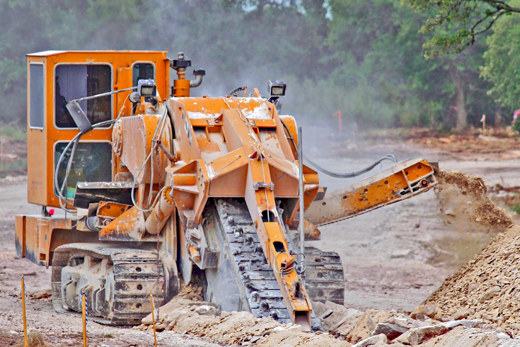 A yellow construction machine grinds and removes earth, creating a pile of dirt on a construction site surrounded by trees and dust.