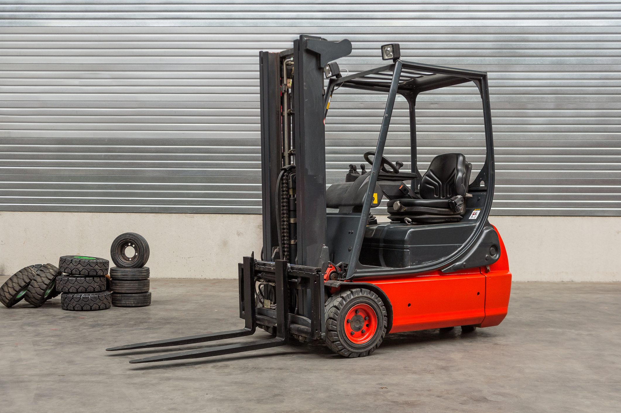 A forklift, positioned stationary with extended forks, rests on a concrete floor beside stacked tires, against a horizontal striped metal backdrop.