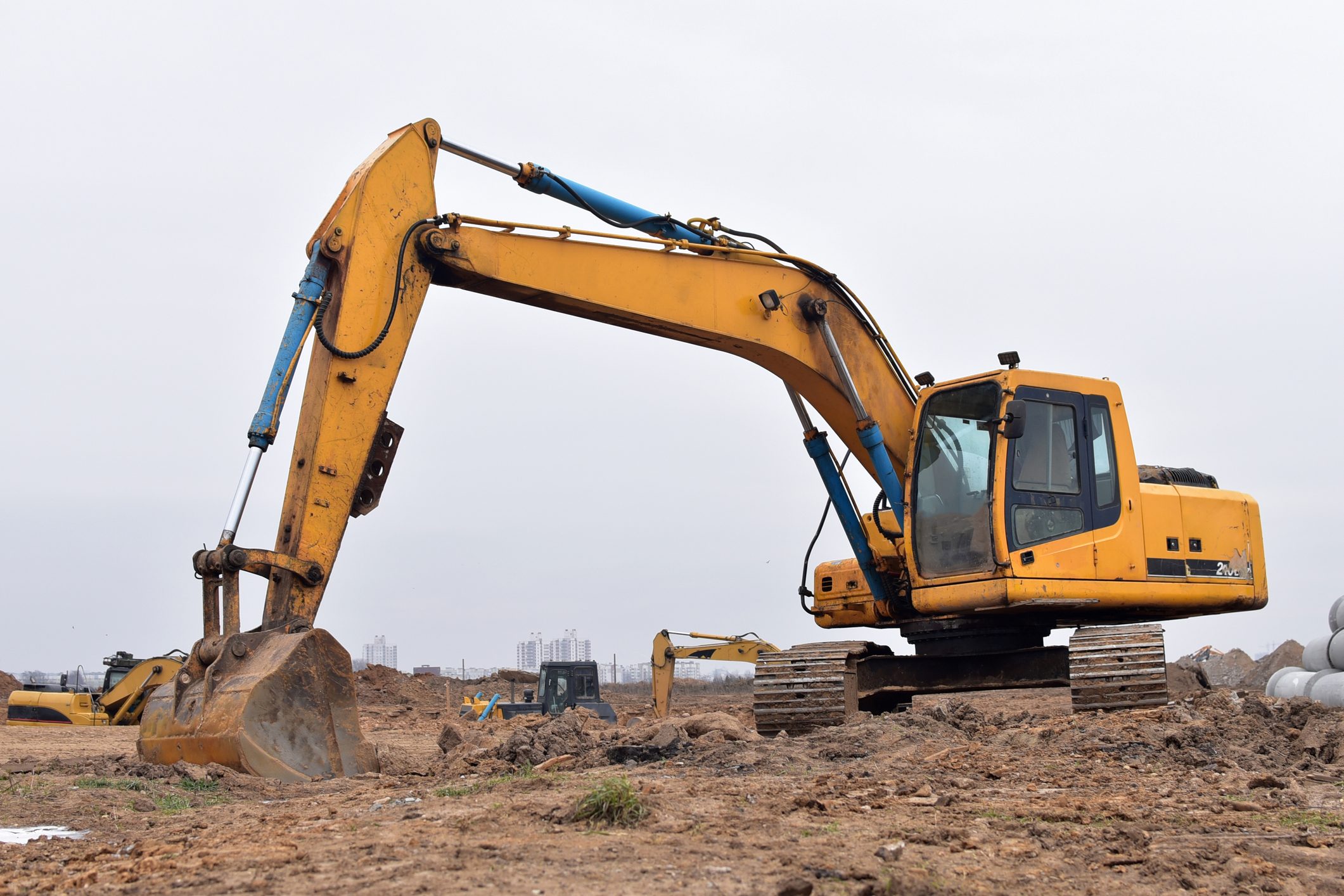 A yellow excavator is positioned on a construction site, its arm extended, ready to dig into the ground amidst dirt mounds and distant machinery.