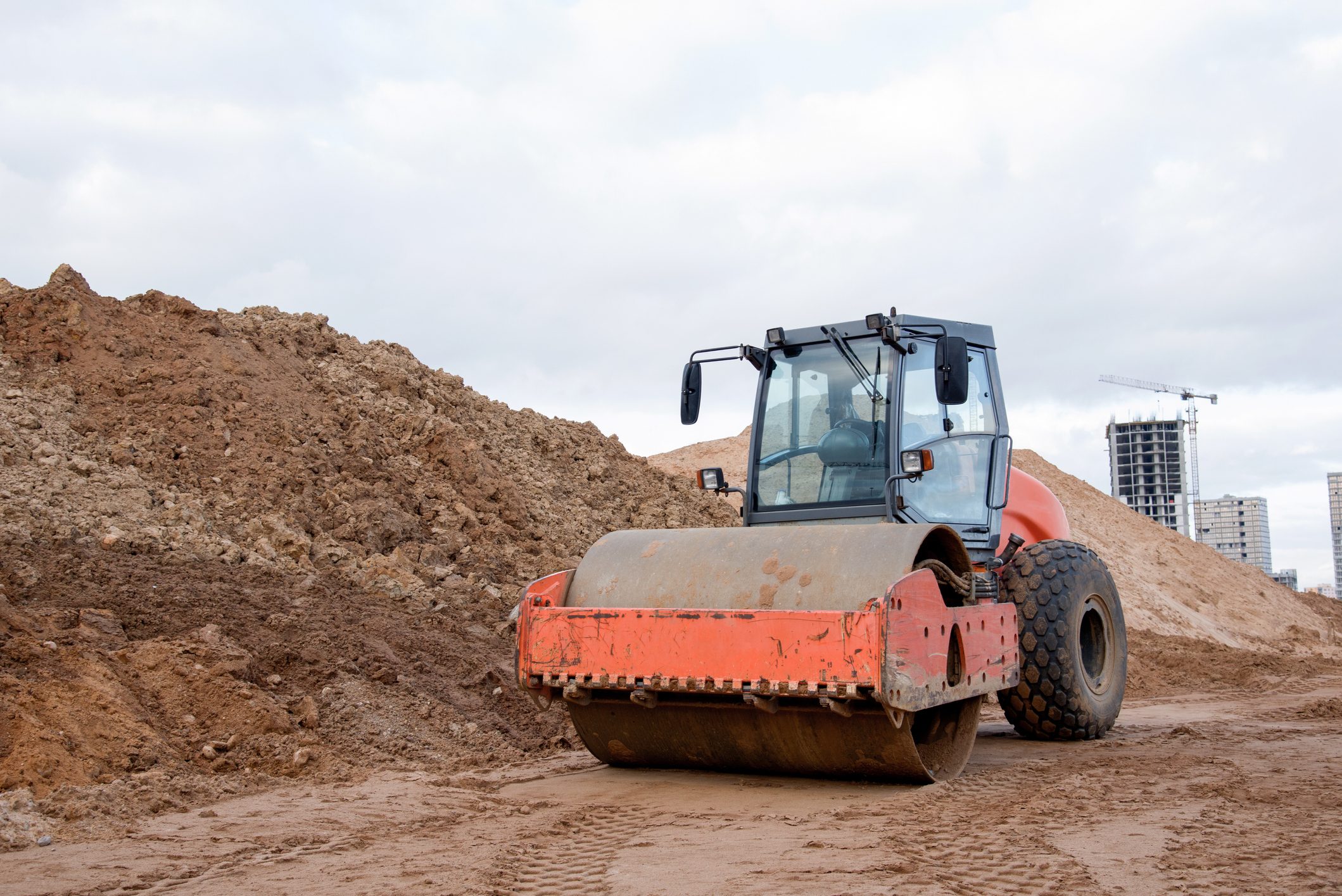 A steamroller compacts earth on a construction site, surrounded by dirt mounds and partially constructed buildings under a cloudy sky.
