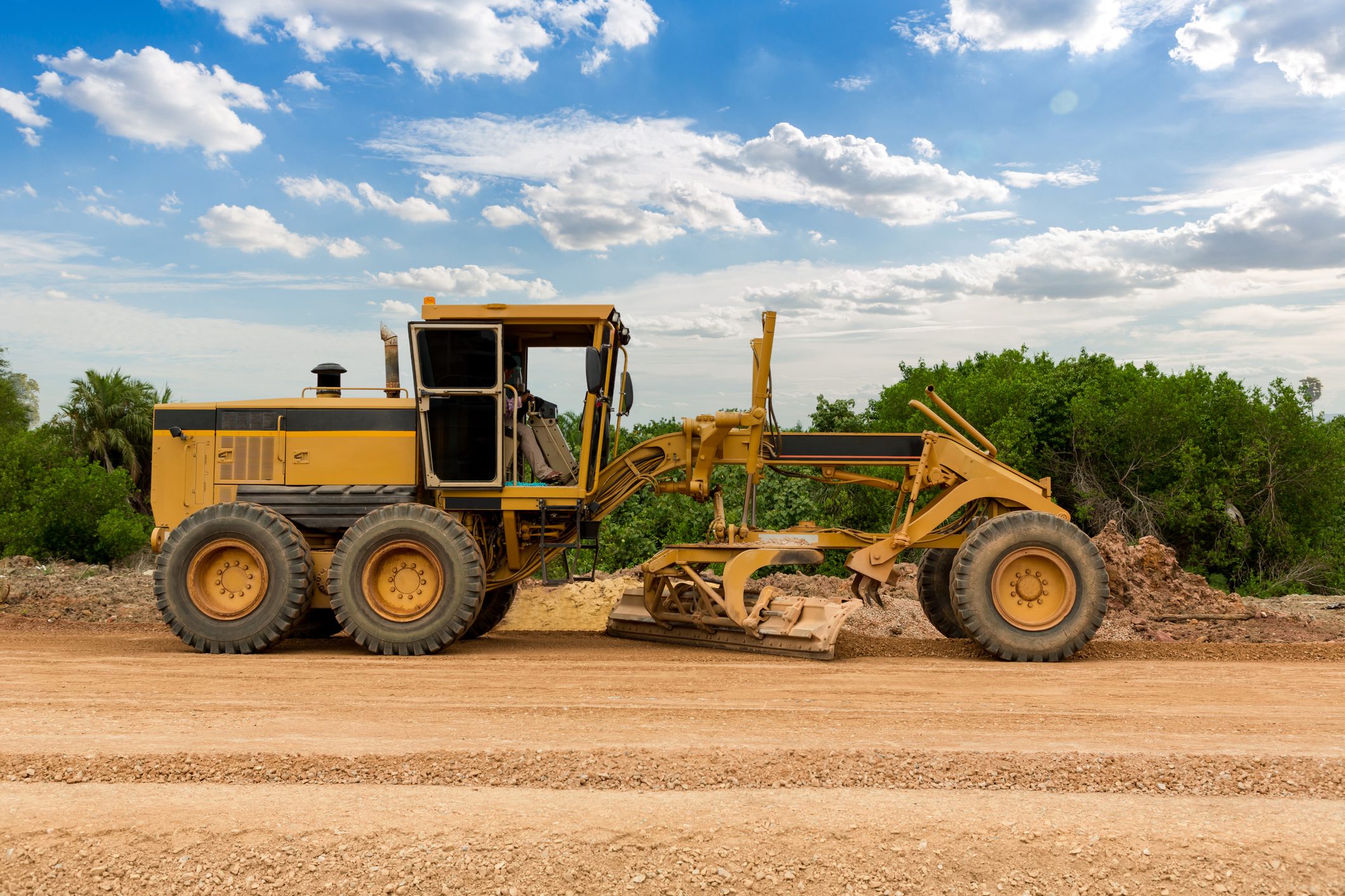 A yellow motor grader levels dirt on a construction site with greenery in the background under a partly cloudy sky.