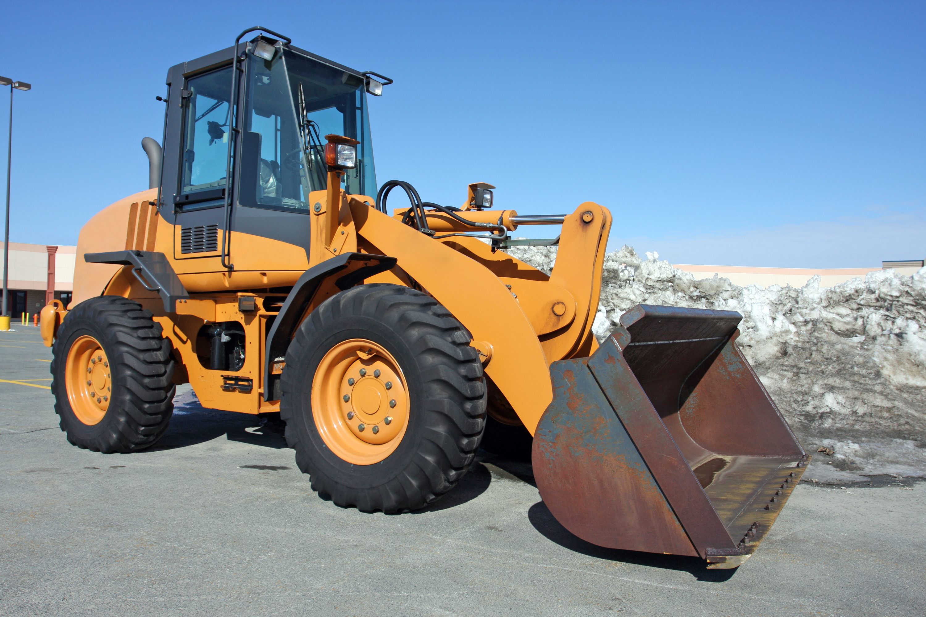 A yellow loader vehicle stands on a parking lot, ready to scoop up snow from a large pile nearby, under a clear blue sky.