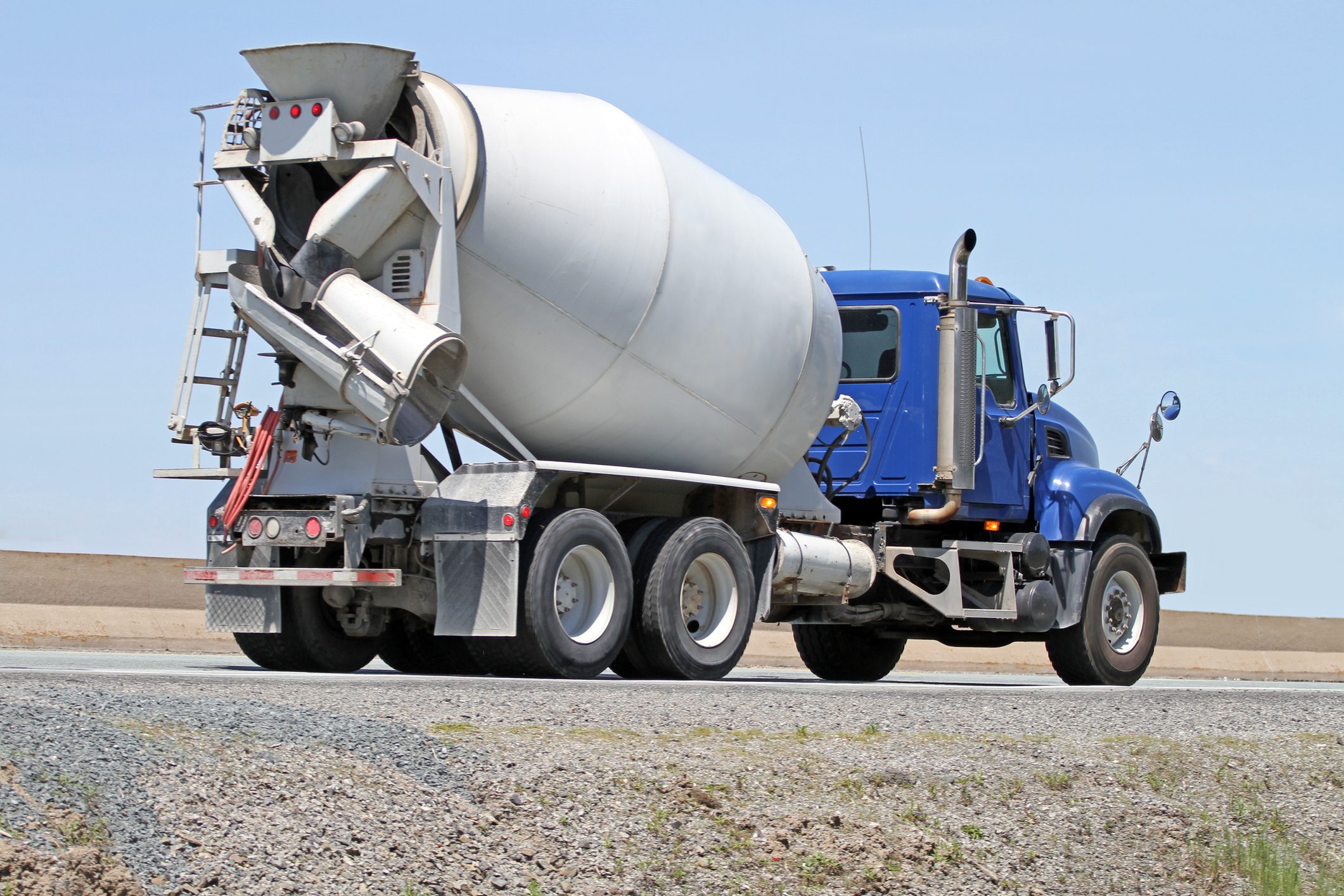 A large concrete mixer truck is parked on a gravel shoulder, under an expansive blue sky, with its drum oriented towards the road.