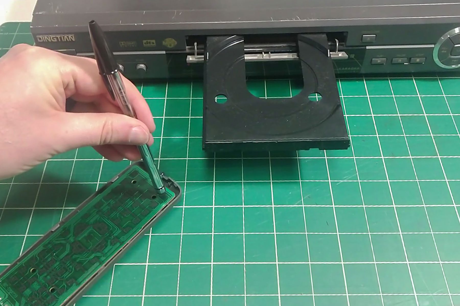A hand holds a pen, tightening screws on a circuit board near an open DVD player, positioned on a green grid cutting mat.