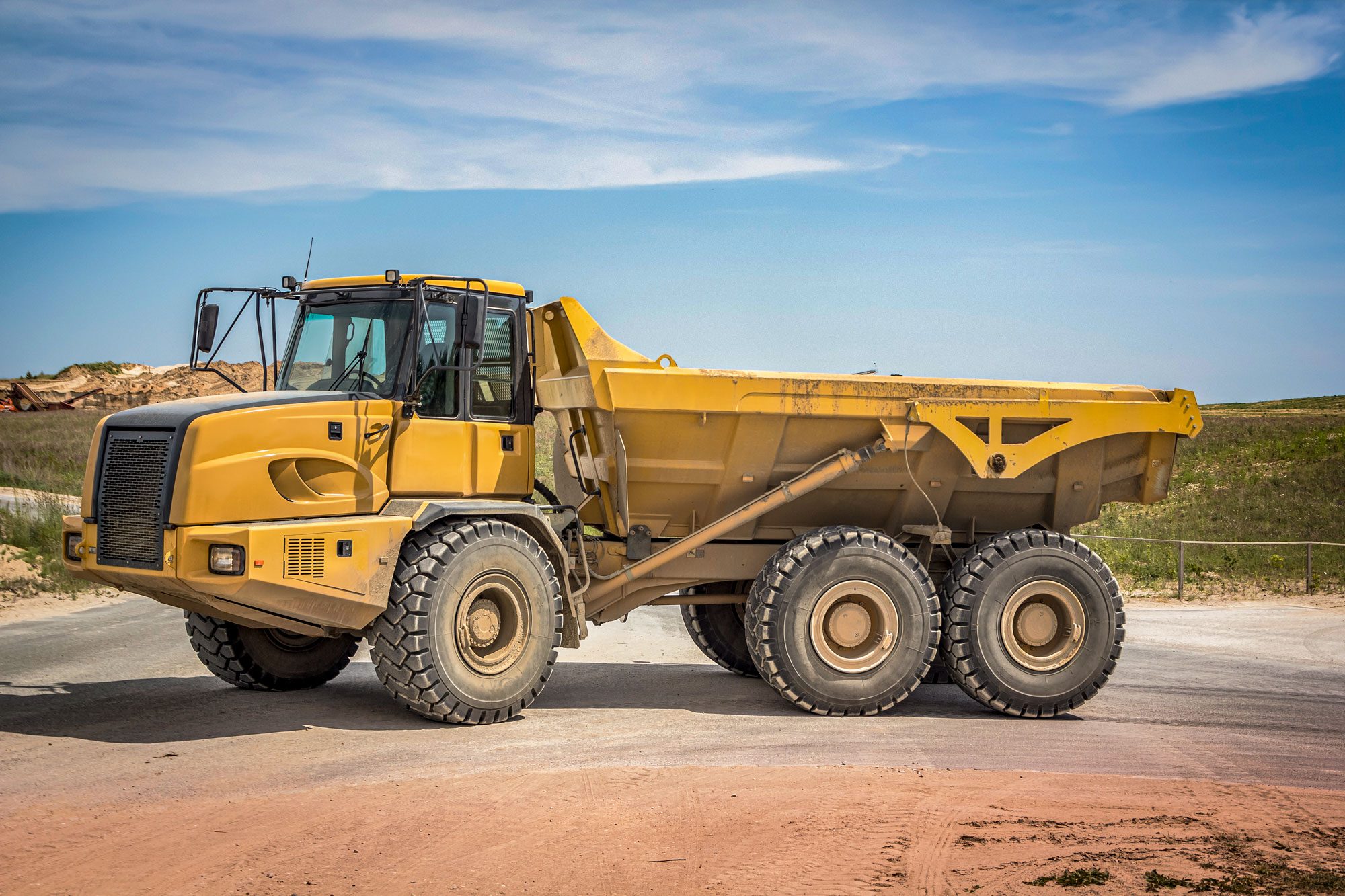 A yellow dump truck stands on a dirt road, surrounded by green fields under a clear blue sky, ready for loading or unloading materials.