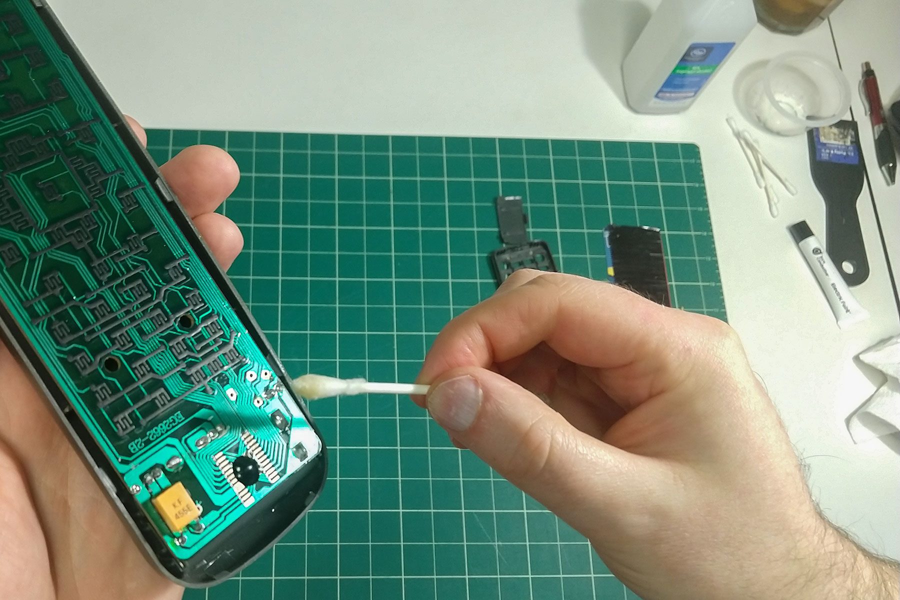A person cleans a circuit board using a cotton swab. They are seated at a green cutting mat with tools and materials scattered around.