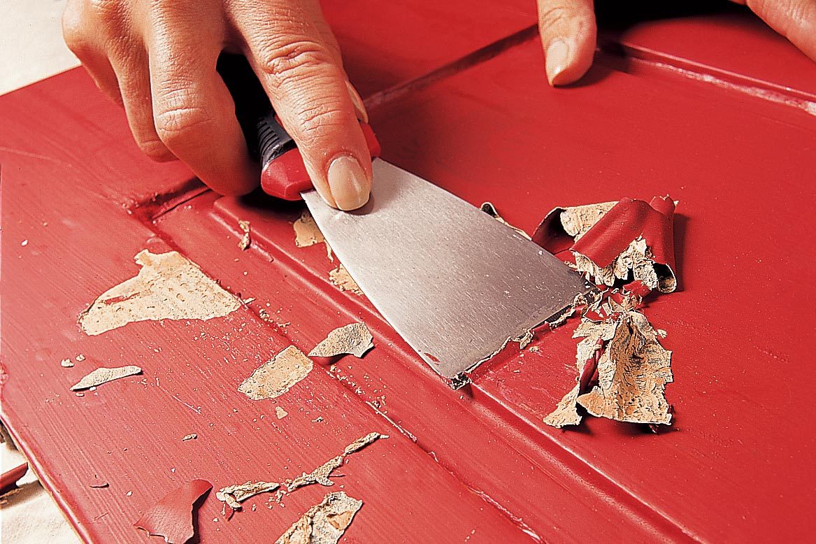 A hand holds a scraper, removing paint from a red surface. Scraps of paint gather as the tool glides over the textured area.