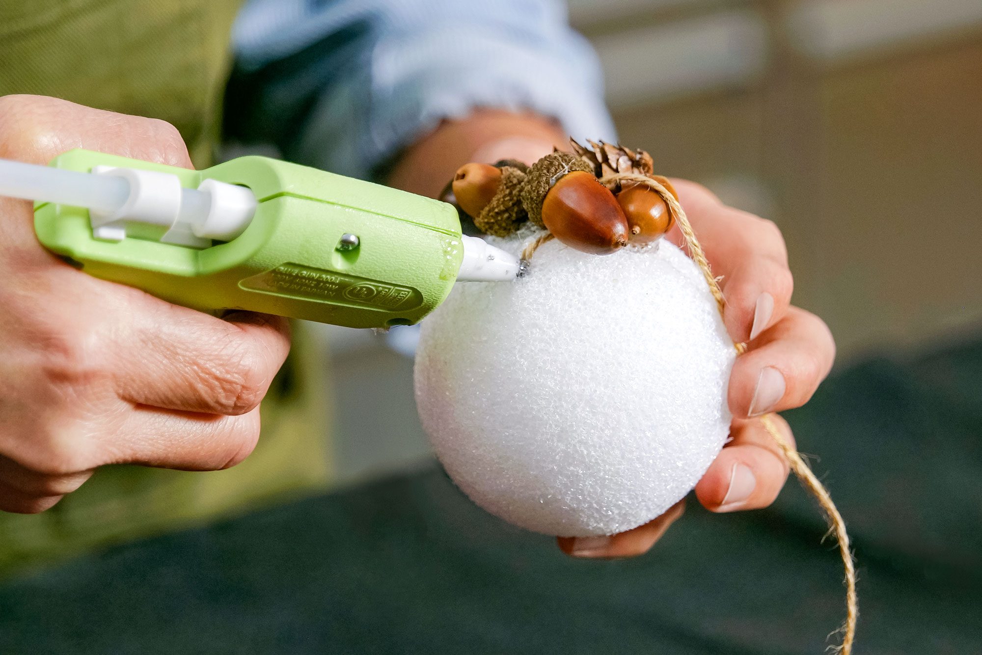 A person uses a green hot glue gun to attach natural elements onto a white foam ball in a softly lit indoor environment.