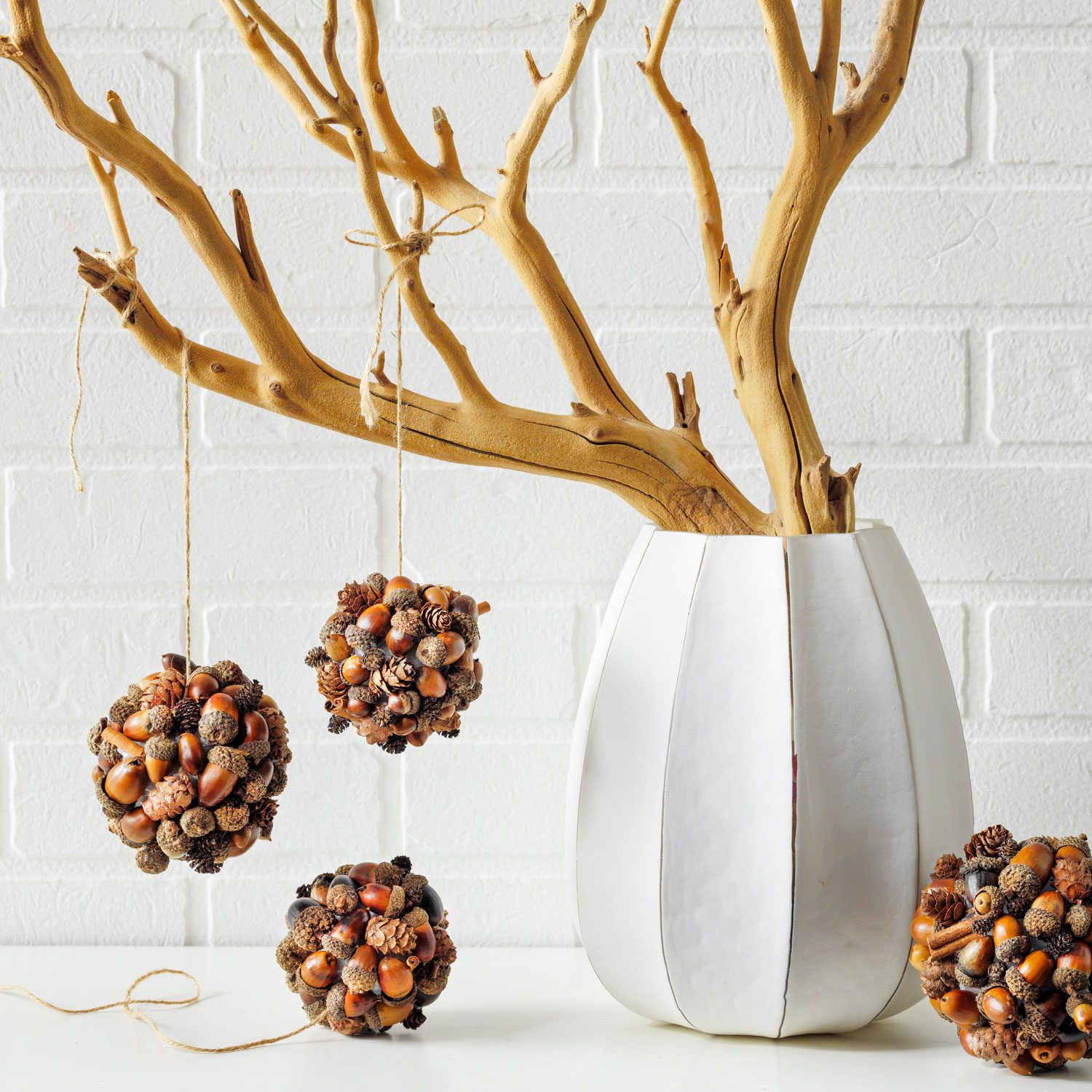 A white vase holds a bare branch, with three decorative balls made of natural materials hanging from twine against a white brick background.