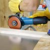 A person wearing yellow gloves uses a blue circular saw to cut a piece of wood secured with clamps on a workbench. The background features a wooden wall and additional tools.