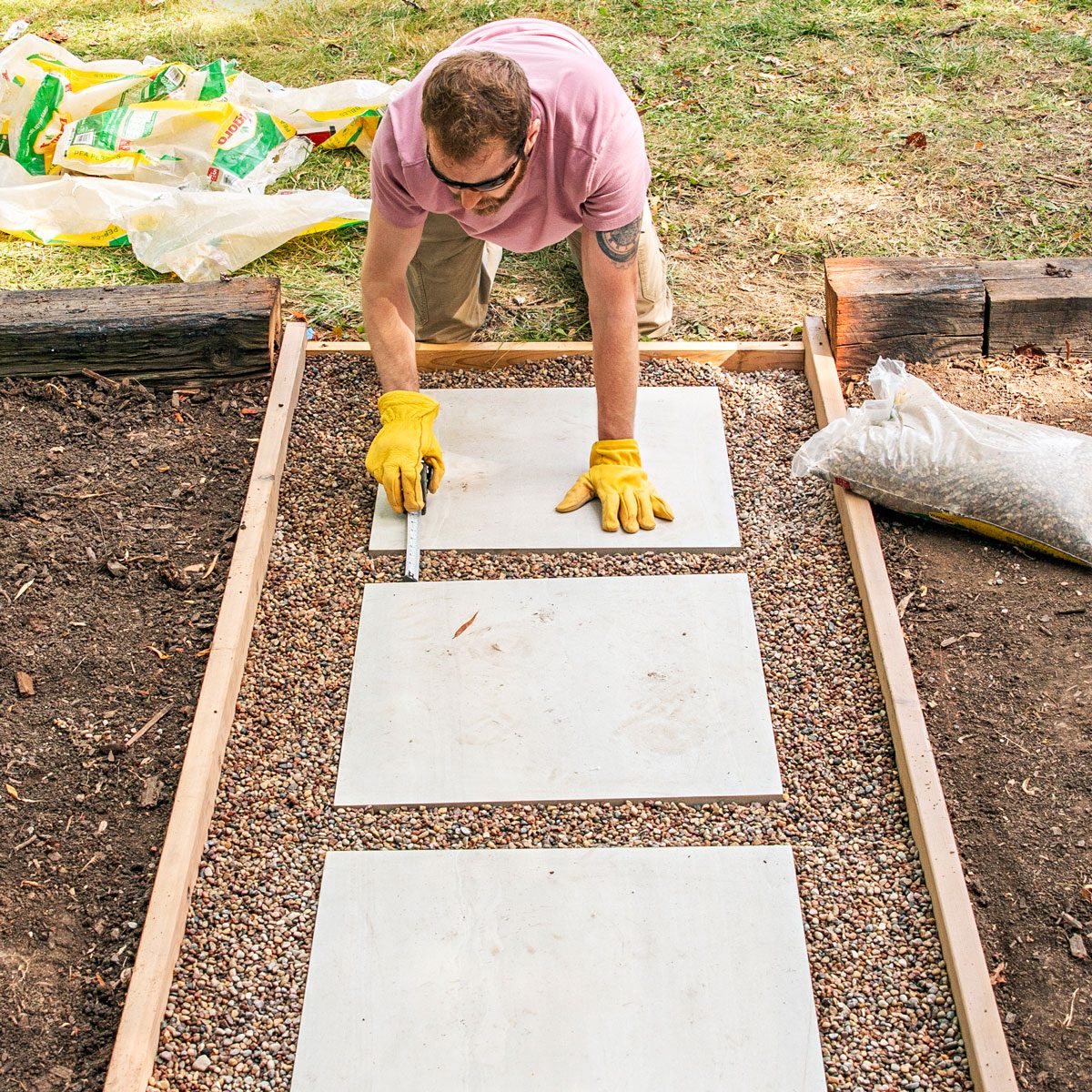a person in a pink shirt and sunglasses carefully installing or aligning white cement boards or pavers in a linear pattern over a gravel-filled wooden frame; they're wearing protective yellow work gloves and using what appears to be a measuring tool to ensure proper placement; three white cement boards are visible in sequence, creating what looks like a pathway or walking surface; the wooden frame is surrounded by grass and soil